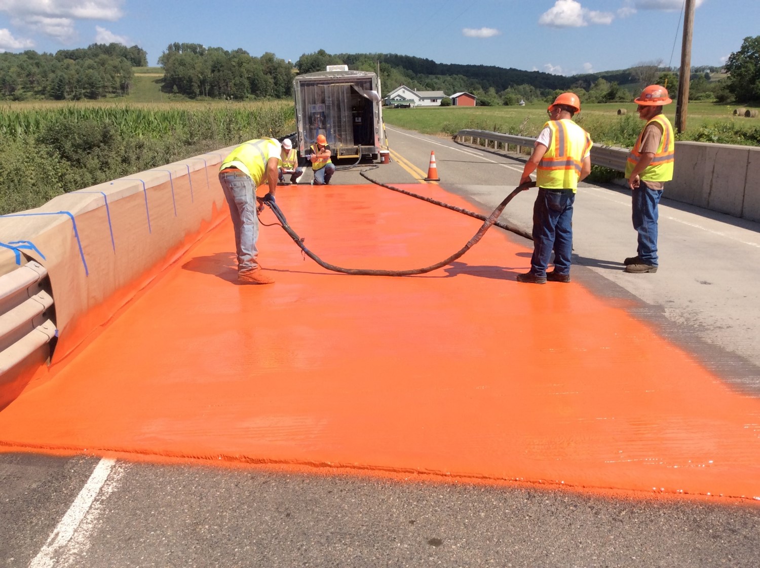 An image of three construction workers in hard hats and yellow safety vests using a sprayer to apply an orange waterproof coating to a bridge deck